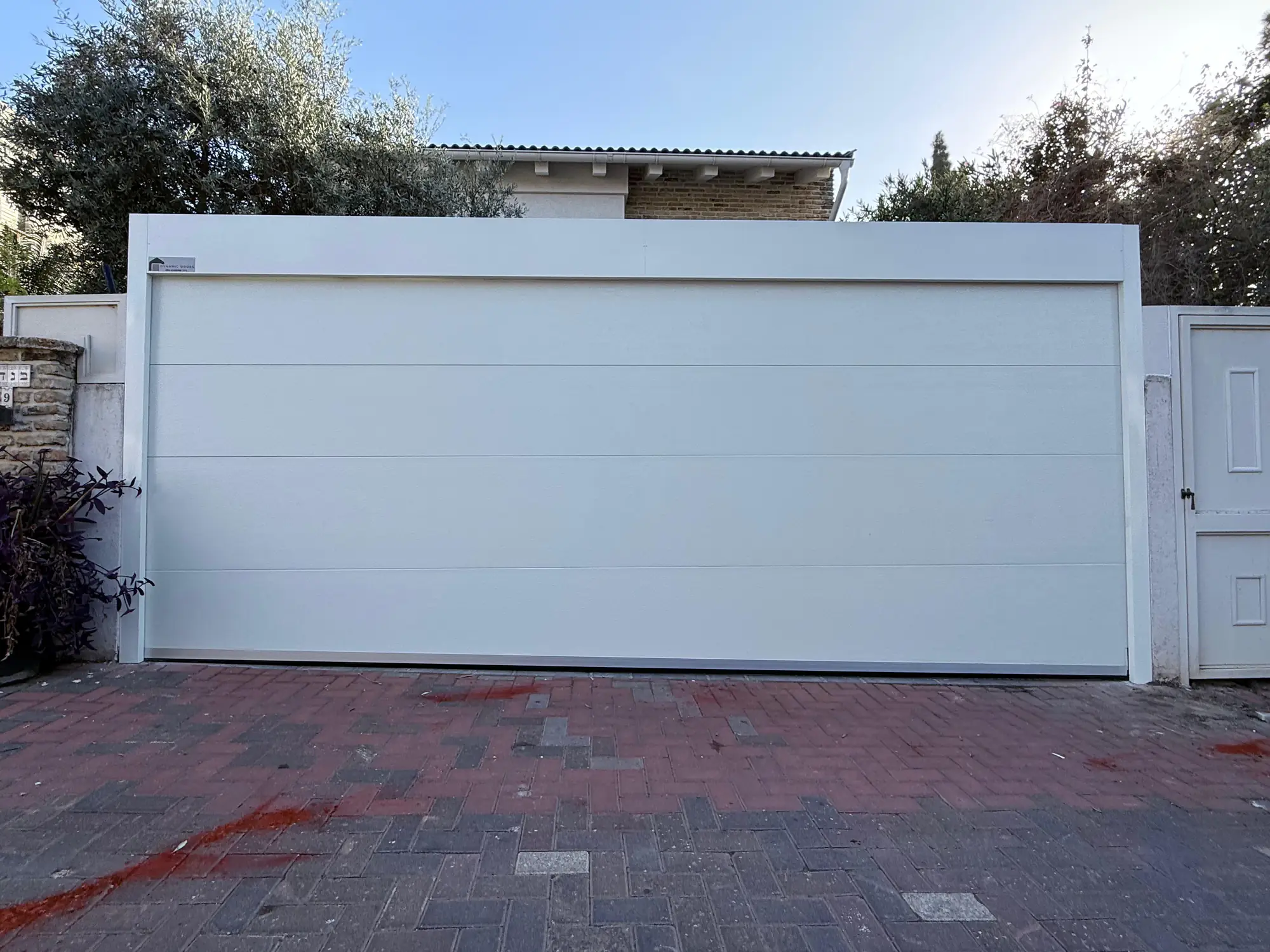 דלת חניה לבנה רחבה עם פנלים אופקיים ומסגרת לבנה לצד שער כניסה תואם